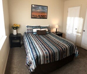 A bedroom with a patterned bedspread, a nightstand with a lamp, a vase with flowers, and a framed picture on the wall. - Cedar Canyon Condos (Blanding)