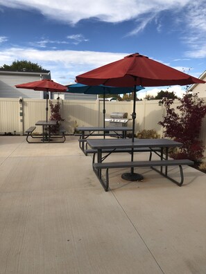 A patio area with picnic tables and red umbrellas, a barbecue grill, and a wooden fence. - Cedar Canyon Condos (Blanding)
