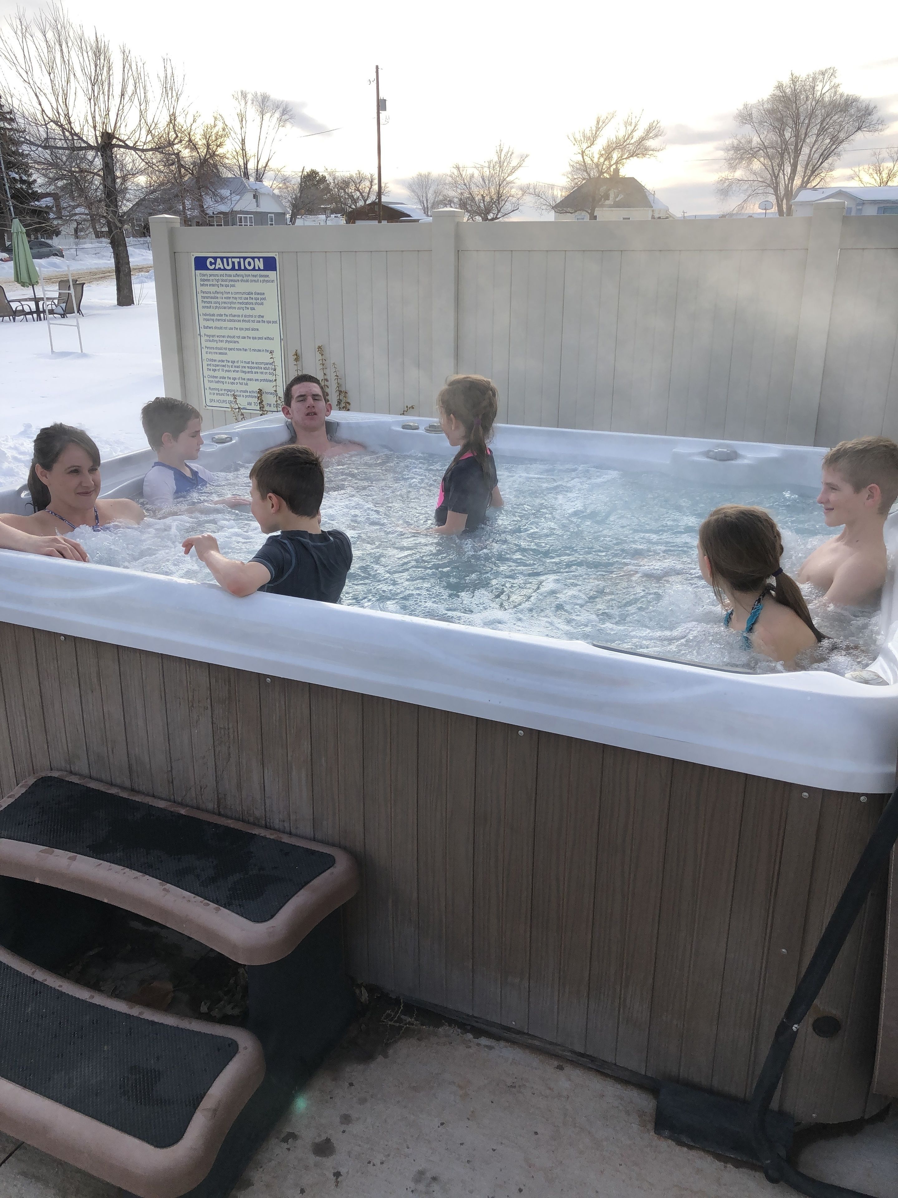 A hot tub with people around it, surrounded by a wooden fence, and a caution sign visible.