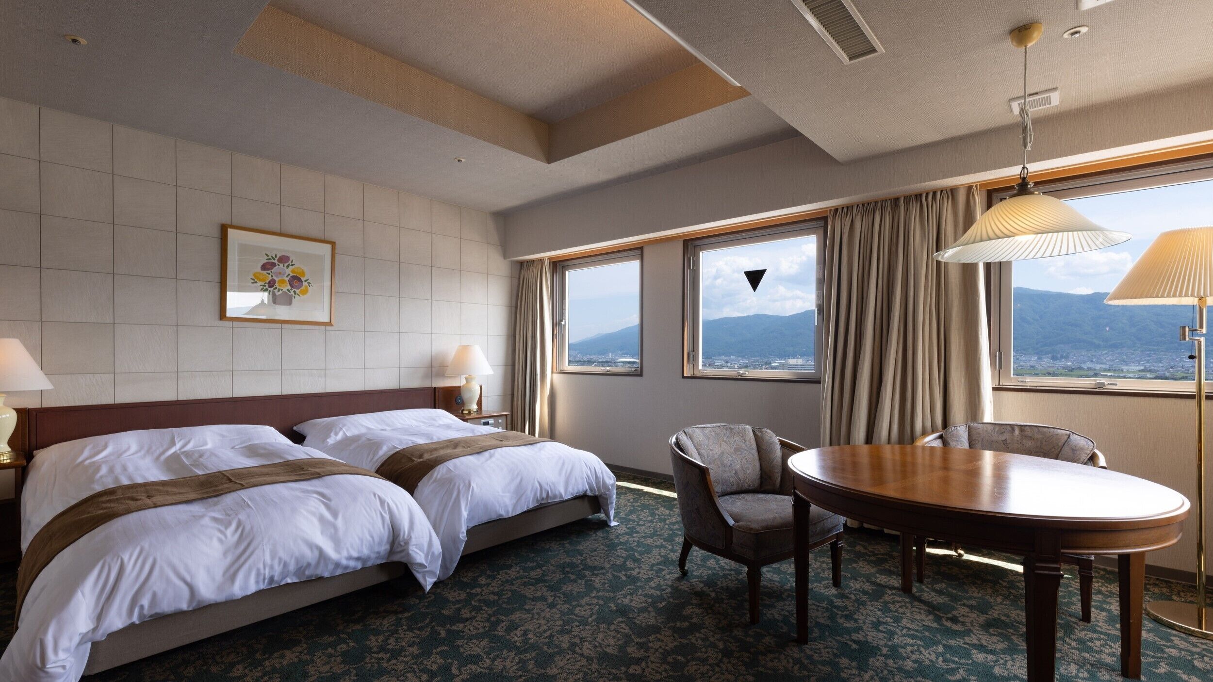 A hotel room with two beds, a dining table, a chair, a lamp, and a view of mountains.