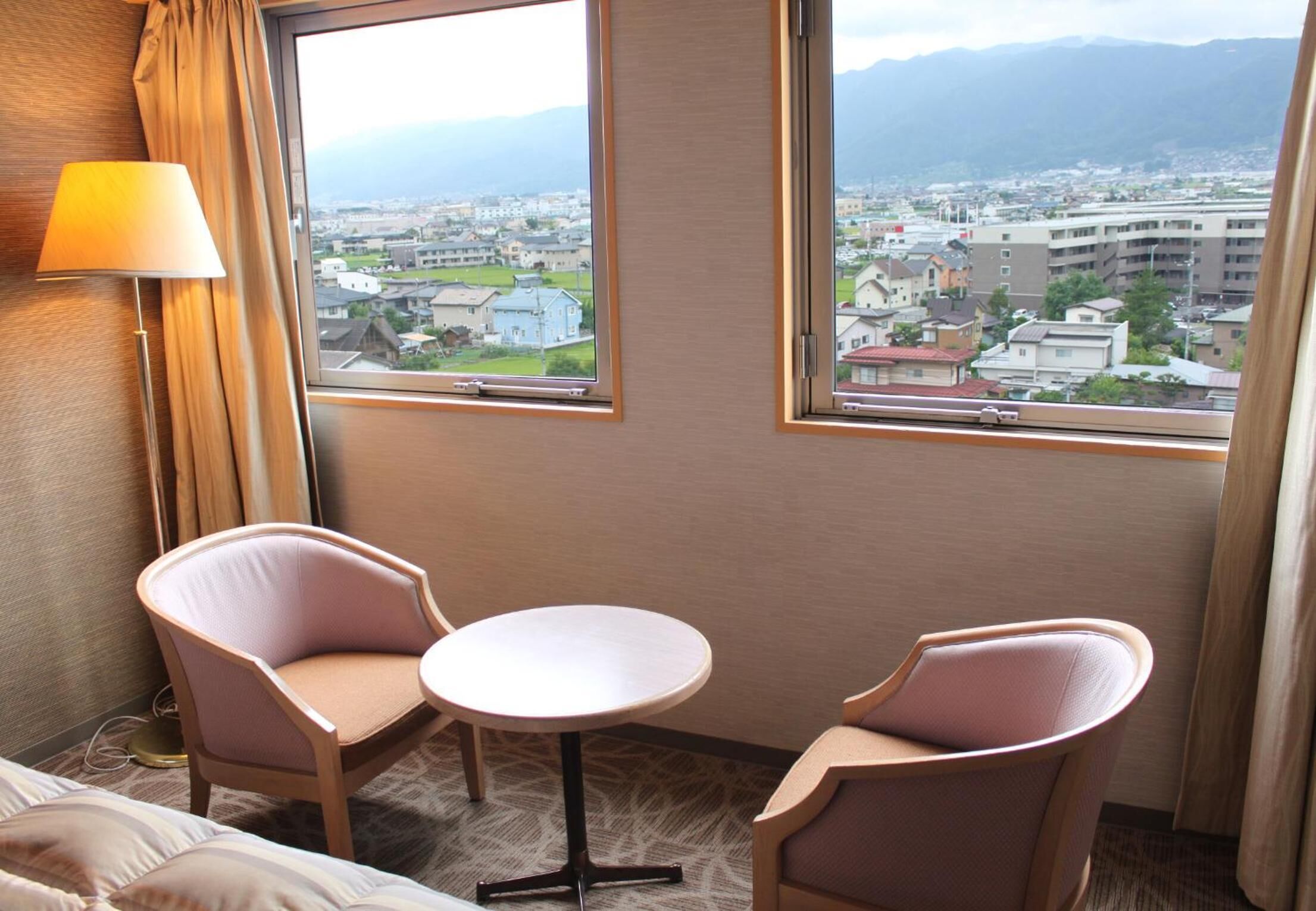 A hotel room with two chairs, a small table, and a view of a cityscape through large windows.