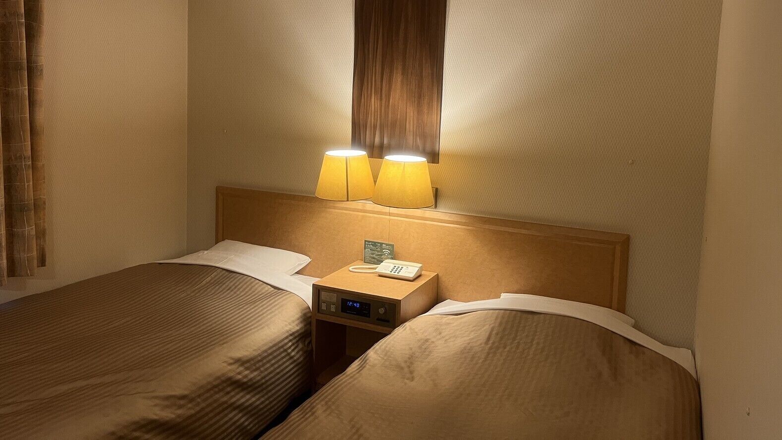 A hotel room with two beds, a nightstand with a phone and a digital clock, and two bedside lamps.