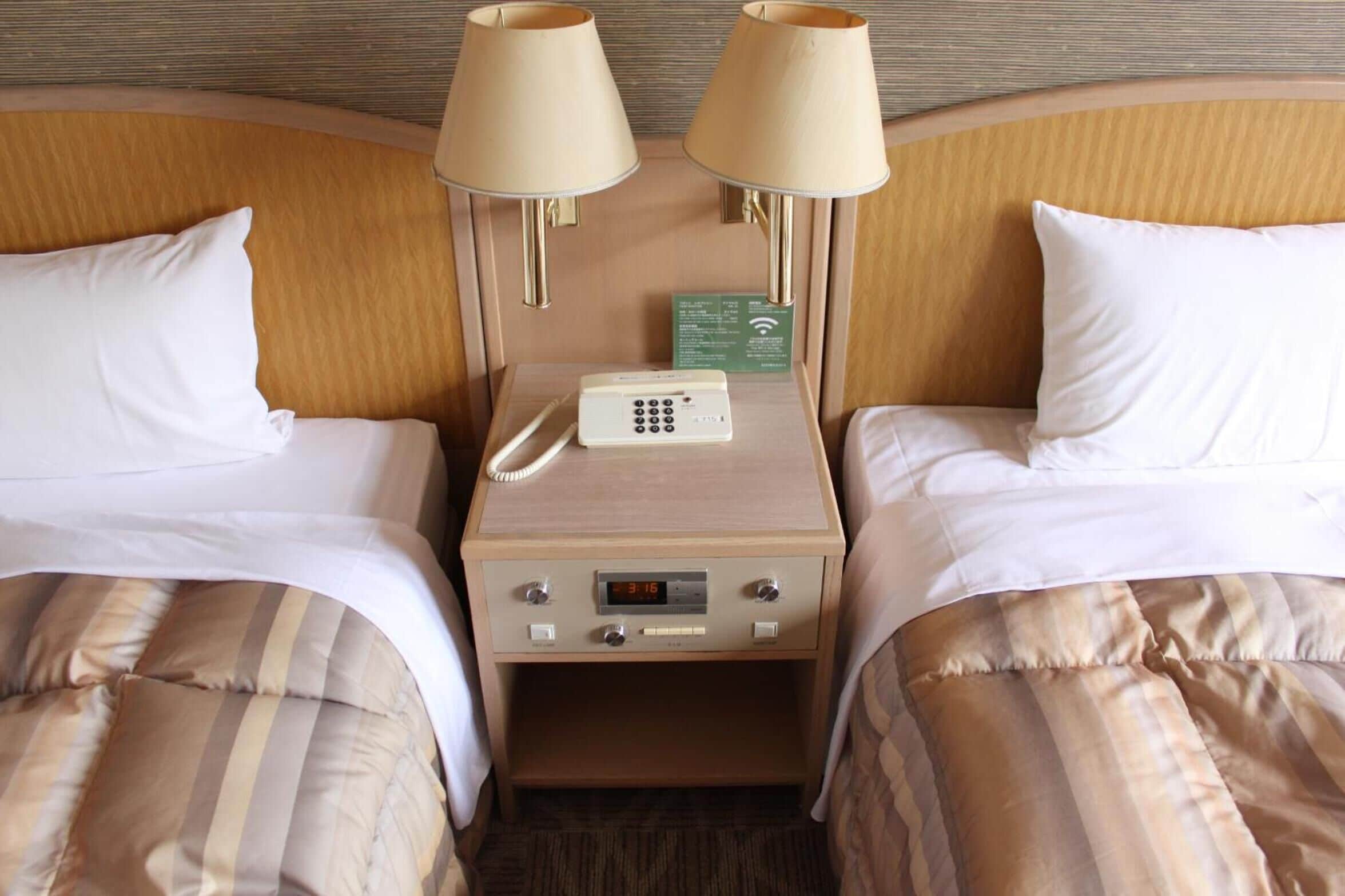 Habitación de hotel con dos camas, cada una con una almohada y una manta a rayas. Hay una mesita de noche con una lámpara, un teléfono y un reloj digital.