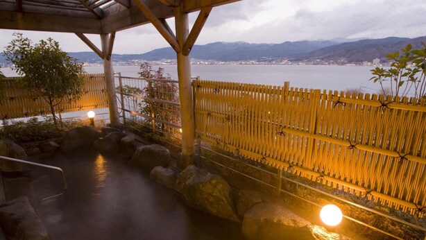 A hot spring area with a wooden fence, rocks, and a view of a lake and mountains in the background. - Rako Hananoi Hotel (Suwa)