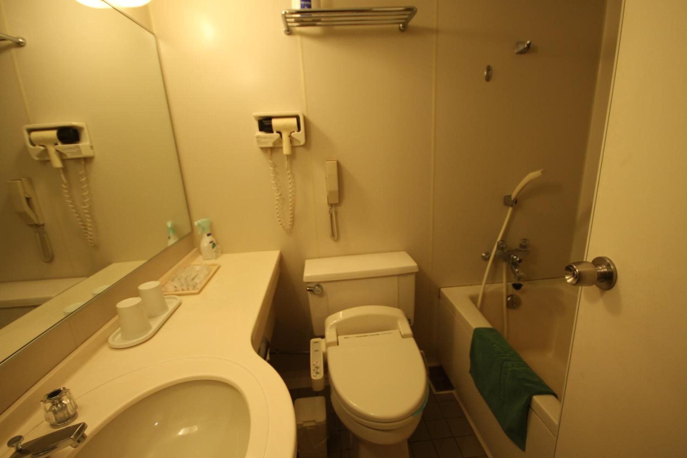 A bathroom with a toilet, sink, and shower. There are two hair dryers mounted on the wall, a towel rack, and a soap dispenser on the sink.