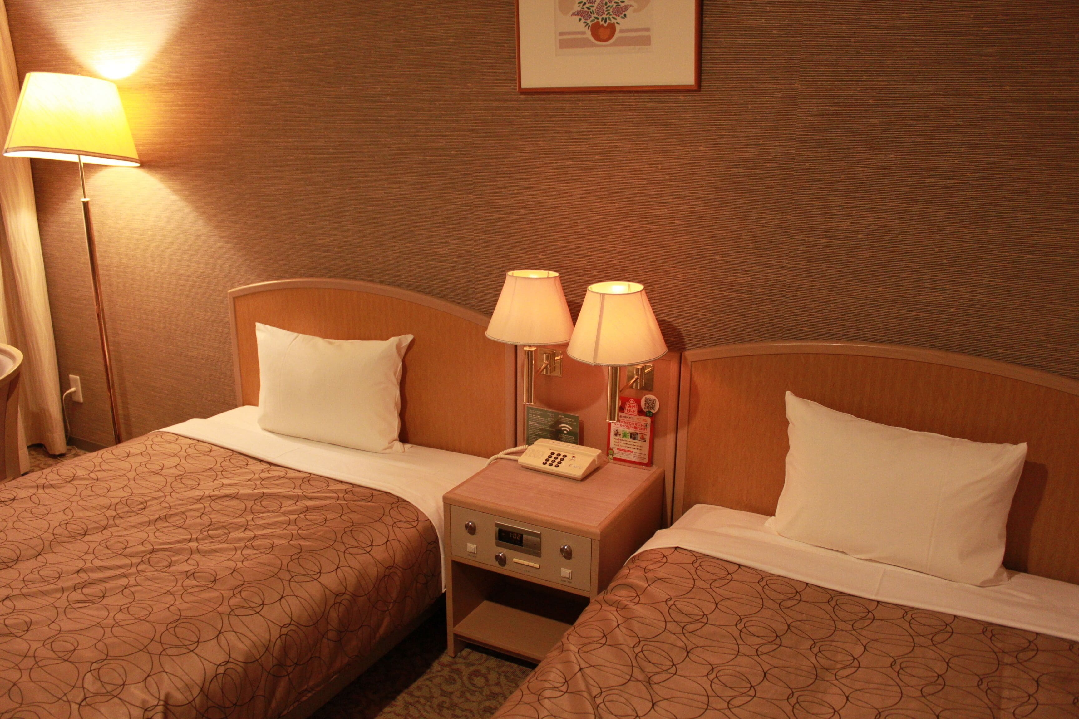 Una habitación de hotel con dos camas, cada una con un edredón estampado, una mesita de noche con una lámpara y una mesita de noche con un teléfono y un reloj.