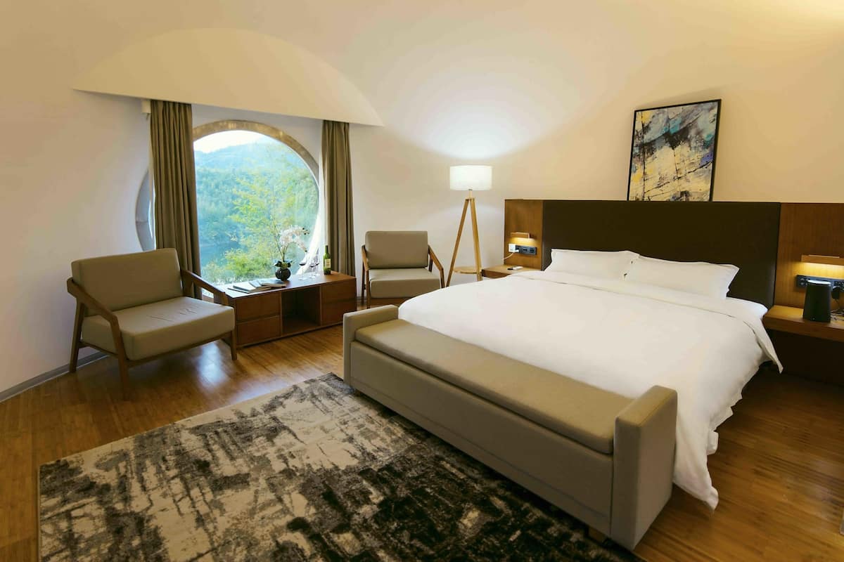Une chambre d’hôtel avec un grand lit, deux fauteuils, une lampe sur pied, une table de chevet et une vue sur l’extérieur grâce à une grande fenêtre.