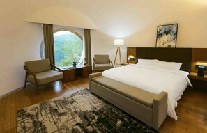 Habitación de hotel con una cama grande, dos sillones, una lámpara de pie, una mesita de noche y vistas al exterior a través de un amplio ventanal.