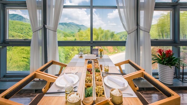A dining table set with various dishes and utensils, large windows with curtains, and a view of green hills. - Planet Zero Retreat (Huzhou)