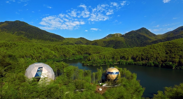 Two geodesic domes blend seamlessly into the mountainous landscape, surrounded by lush greenery and a serene lake.