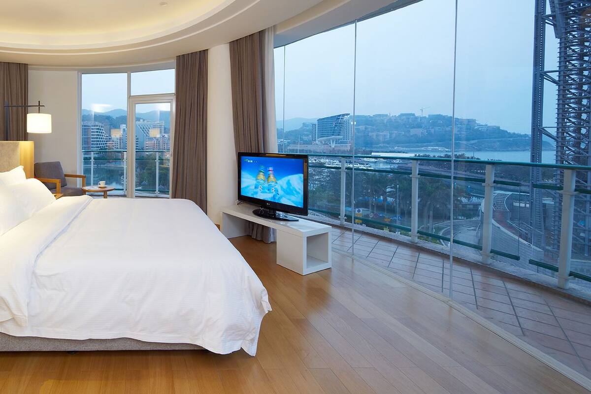 A modern hotel room with a large bed, a television, and a balcony with a city view.