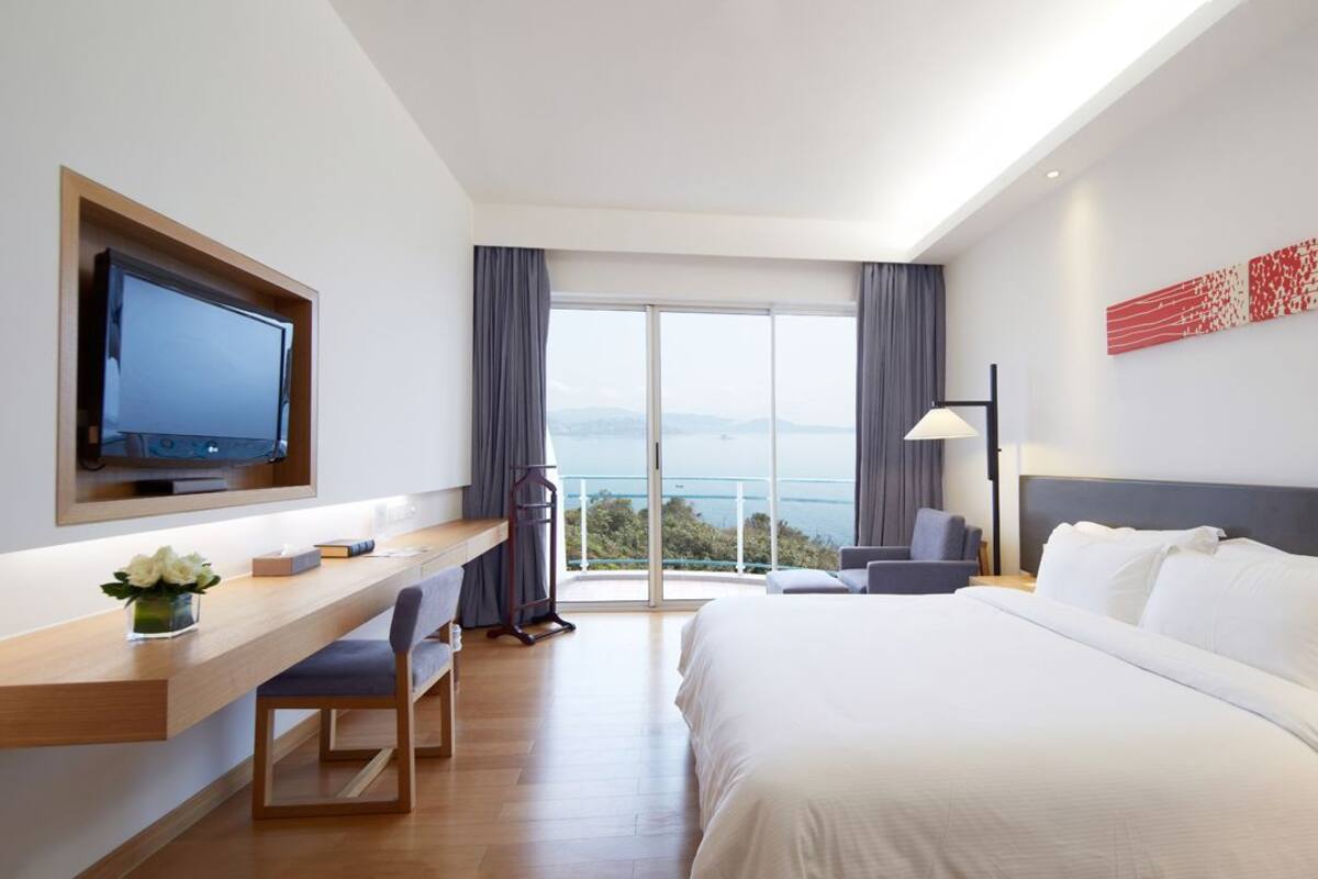 A modern hotel room with a large bed, a desk with a flat-screen TV, a view of the sea, and a red abstract art piece on the wall.