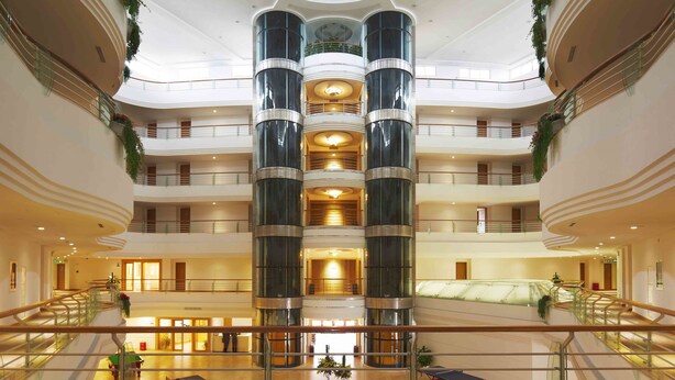 Een meerlaags atrium met een centrale kolom, glazen balustrades en een hoog plafond met een rond dakvenster.