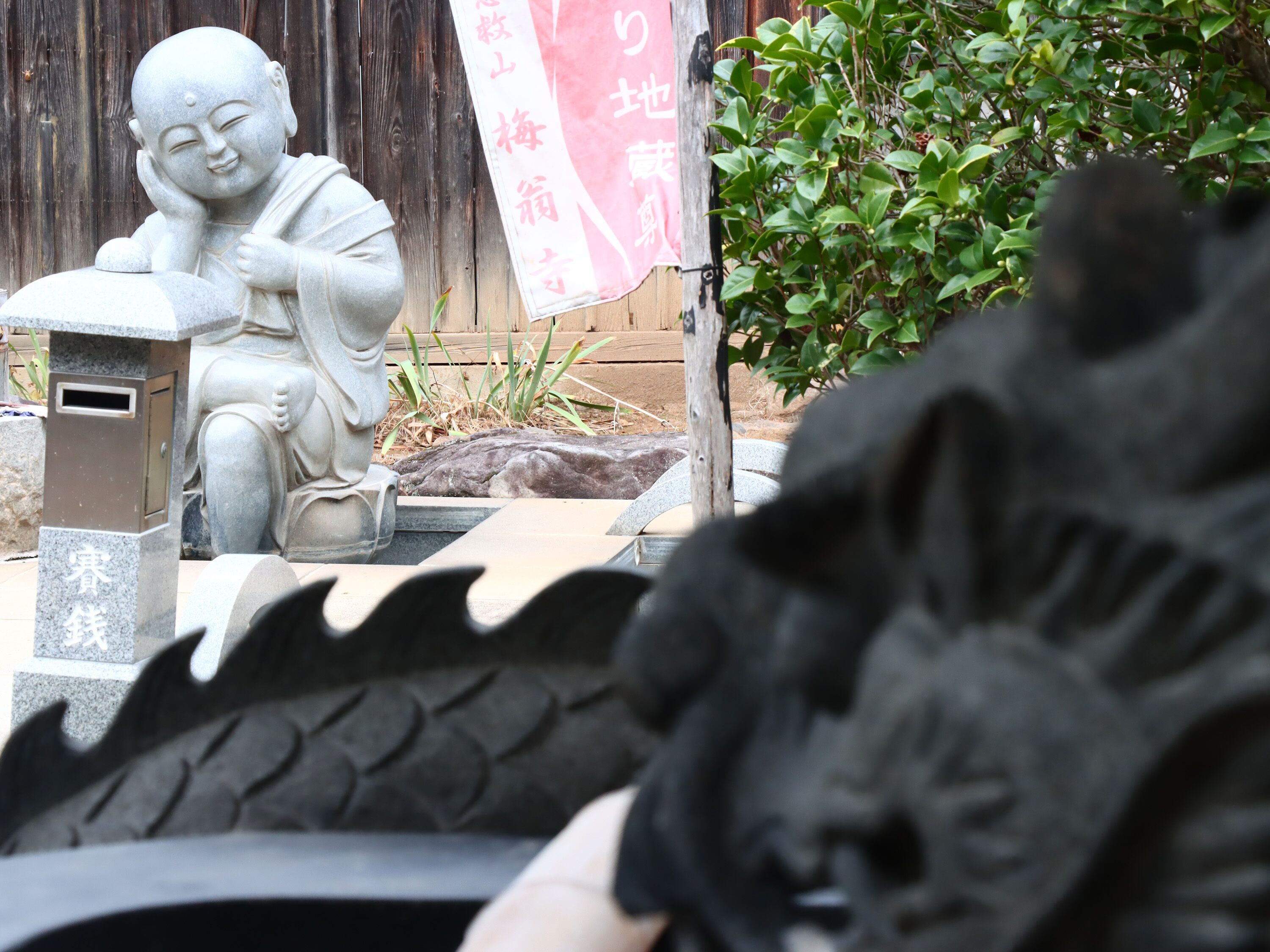 A stone Buddha statue in a garden setting with a wooden fence and greenery in the background.