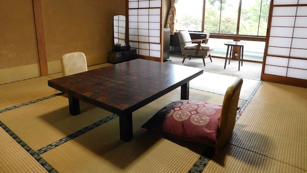 A traditional Japanese-style room with tatami flooring, a low table, and a cushioned chair. - Maruka Ryokan (Yamanouchi)