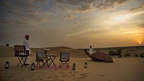 A desert scene with a camel, a man in traditional attire, and a table set with bottles and glasses. - SUJÁN The Serai, Jaisalmer - Relais & Chateaux (Jaisalmer)