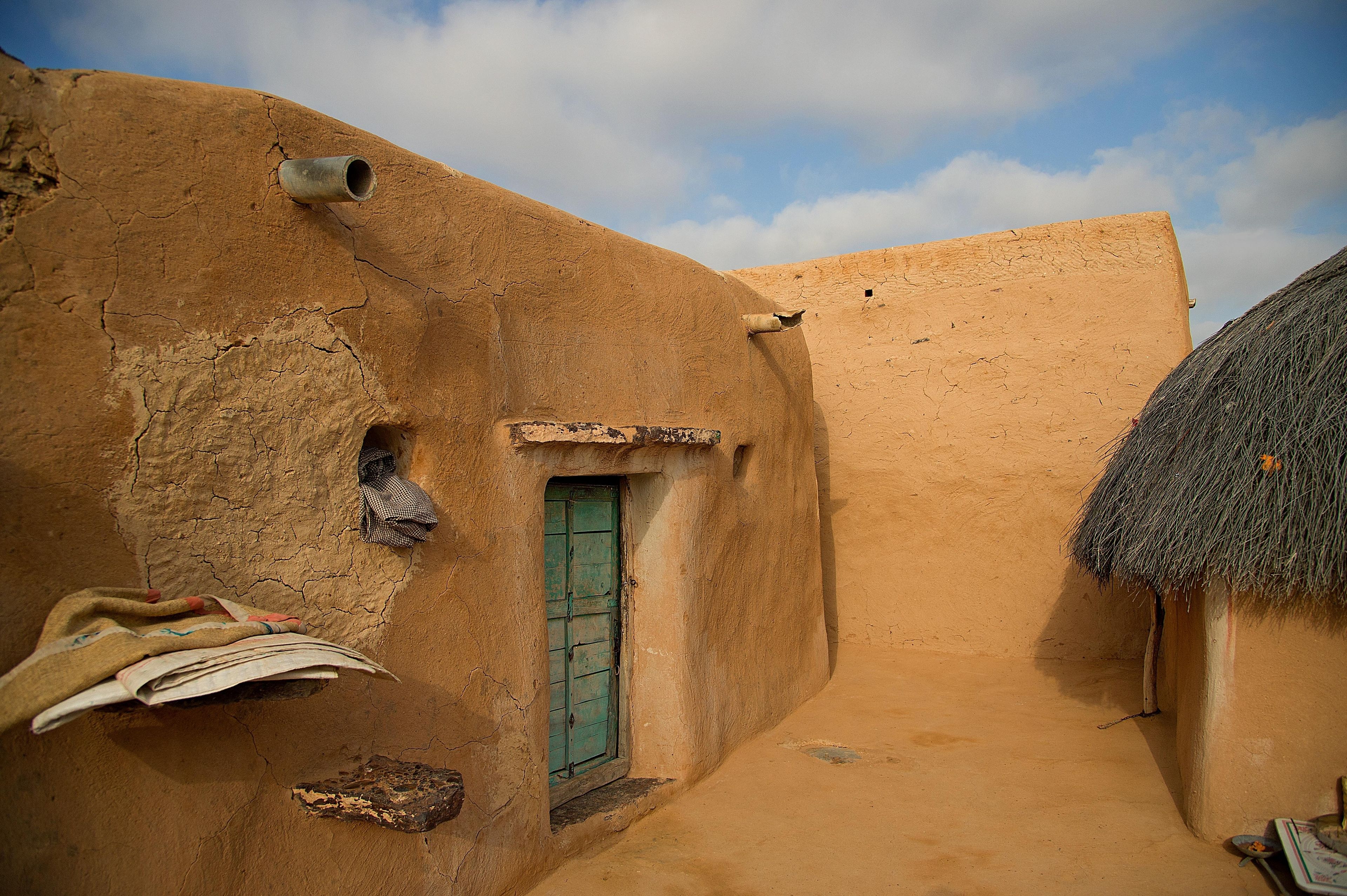Une ruelle étroite bordée de bâtiments traditionnels en briques de terre, une structure à toit de chaume et une petite fenêtre avec une porte verte.