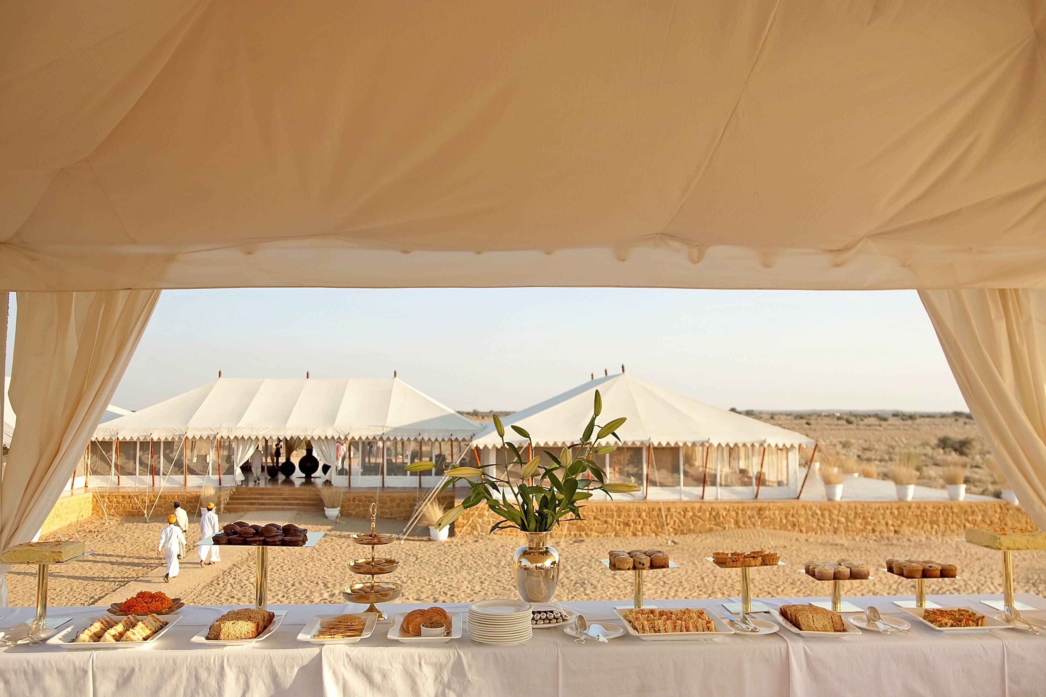 Una mesa de buffet con diferentes comidas y un jarrón de flores, con telones de fondo de tiendas de campaña y un paisaje desértico.