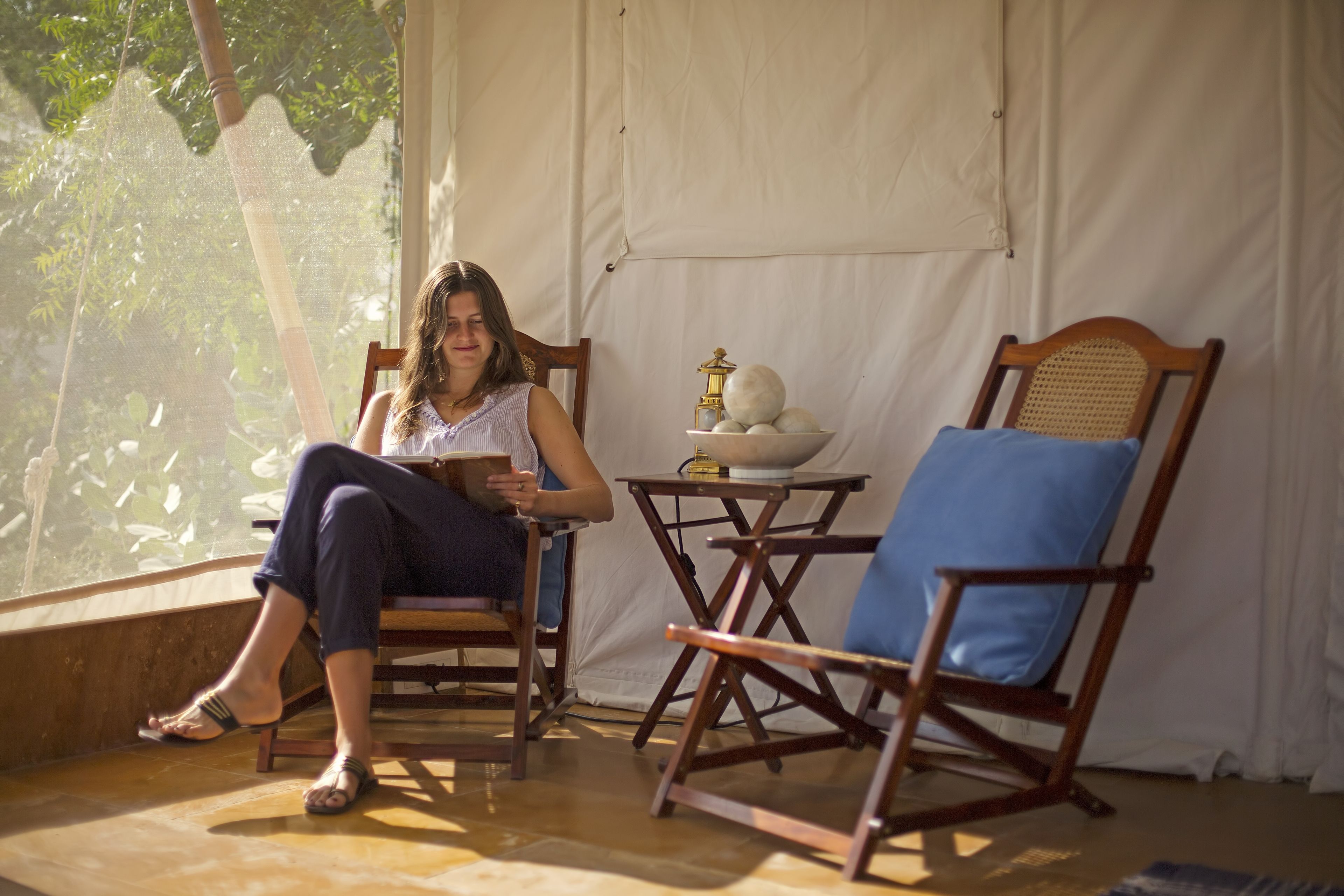 Une personne est assise sur une chaise en bois, dotée d’un coussin bleu, et tient un livre. Elle se trouve dans une pièce avec une grande fenêtre, une petite table sur laquelle se trouve une lampe, et un rideau blanc en arrière-plan.