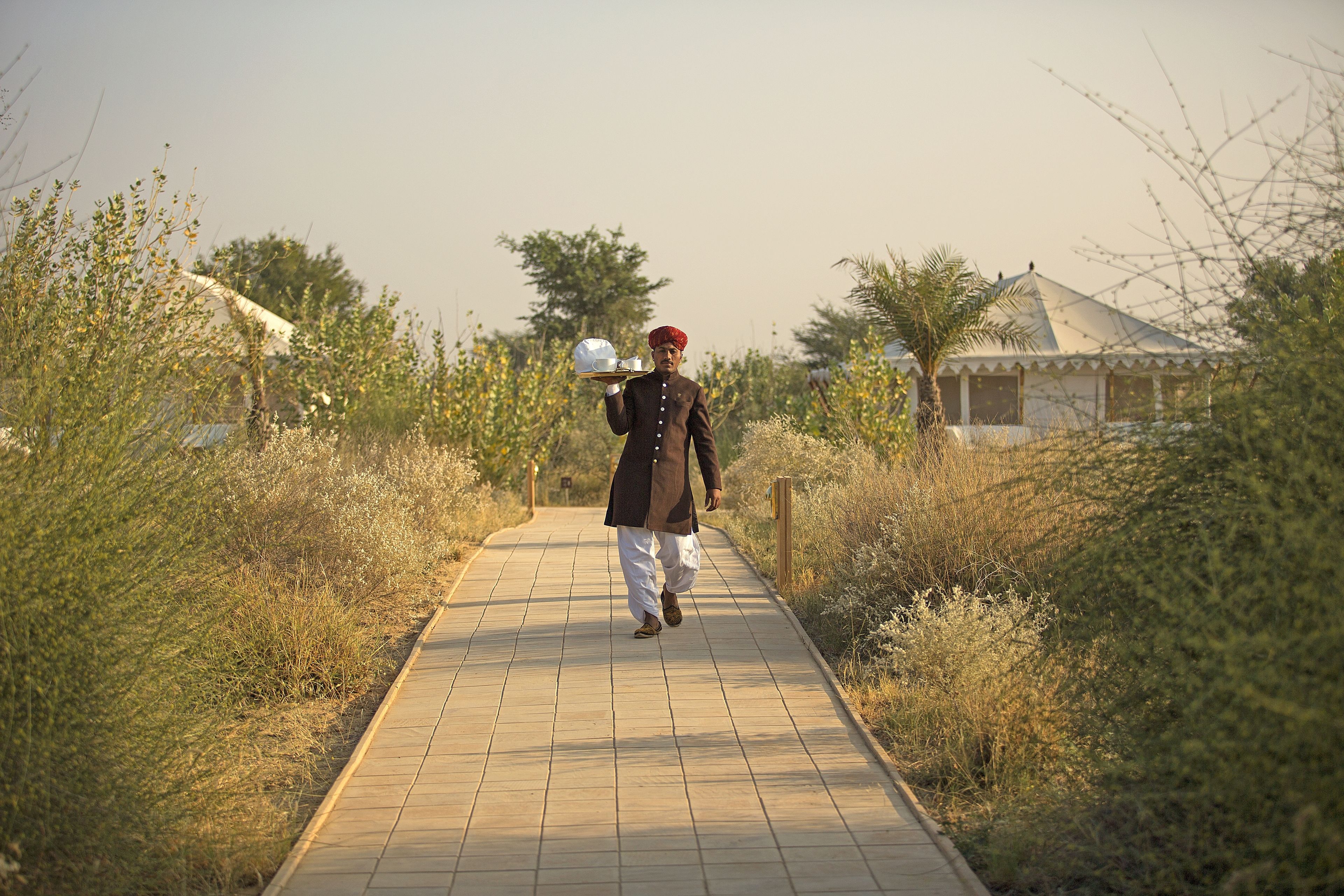 A man walking on a paved pathway surrounded by dry vegetation and a building in the background.