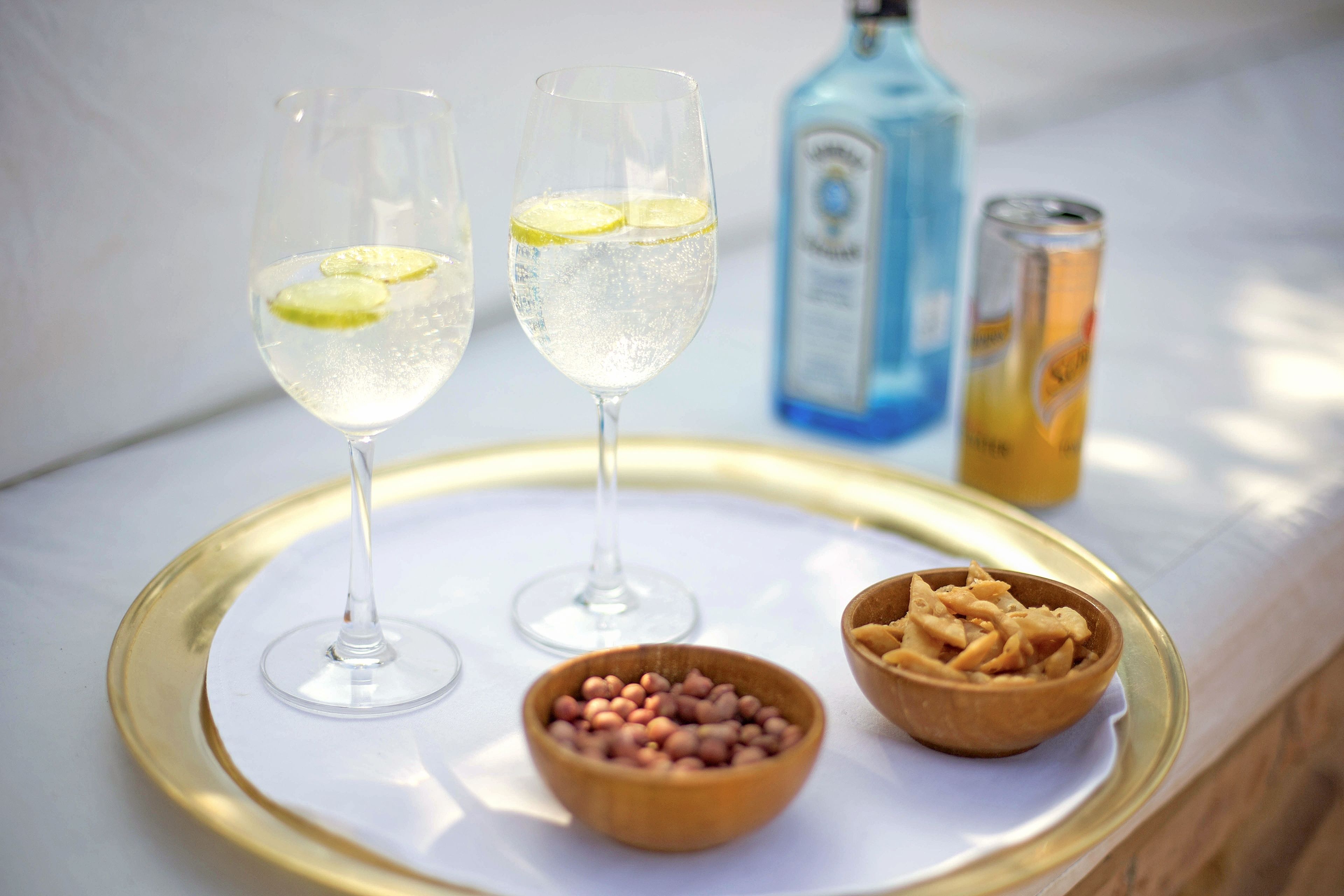 Two glasses of a clear beverage with lime slices, a bottle of blue liquid, a can of yellow liquid, and two bowls of food on a tray.