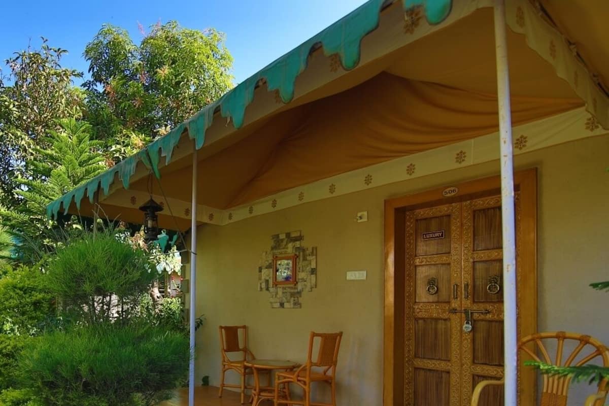 A covered outdoor seating area with two wooden chairs and a table, surrounded by lush greenery.