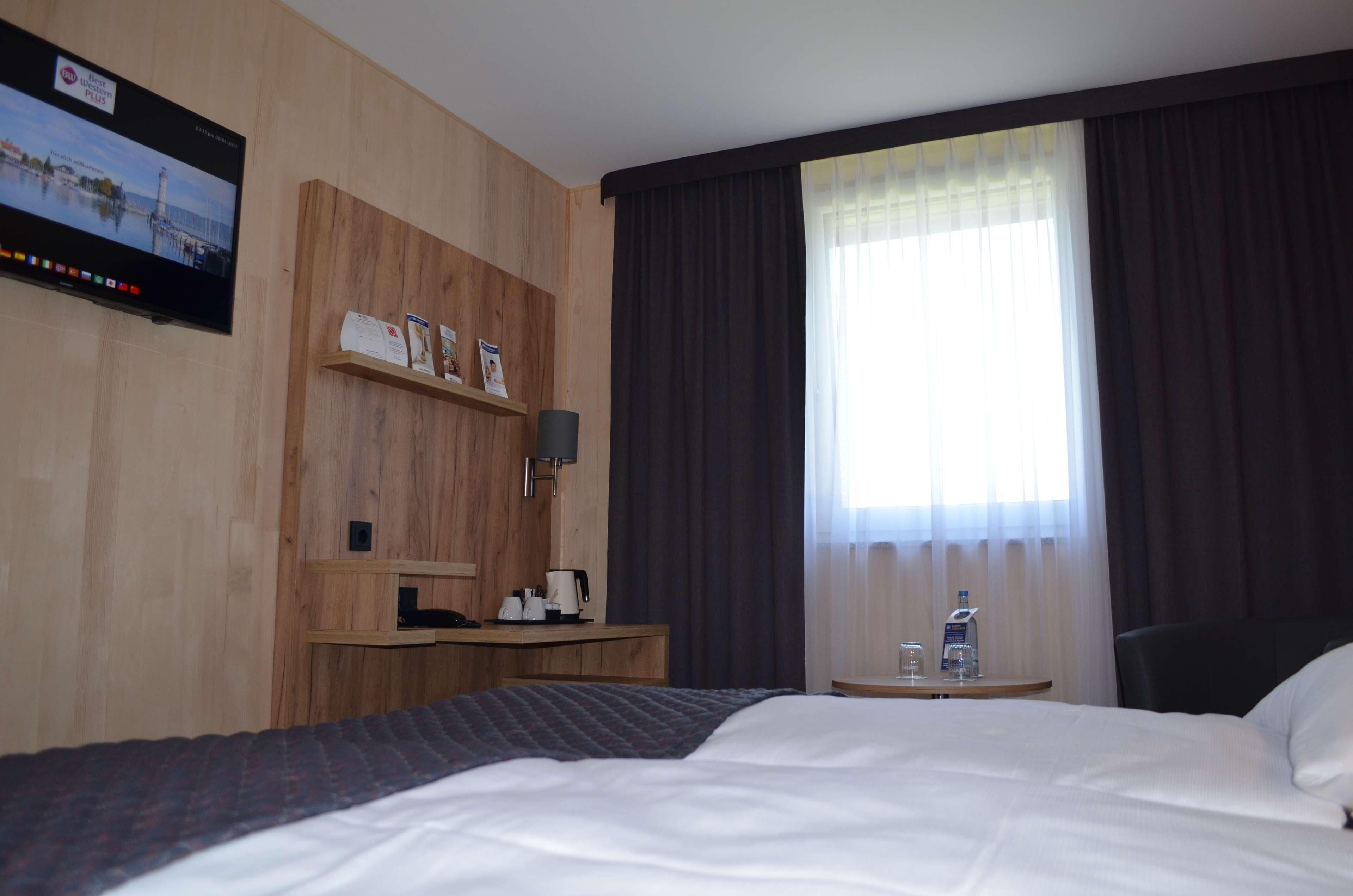 Habitación de hotel con cama, escritorio con lámpara, pequeña mesa con botella de agua y televisión montada en la pared.