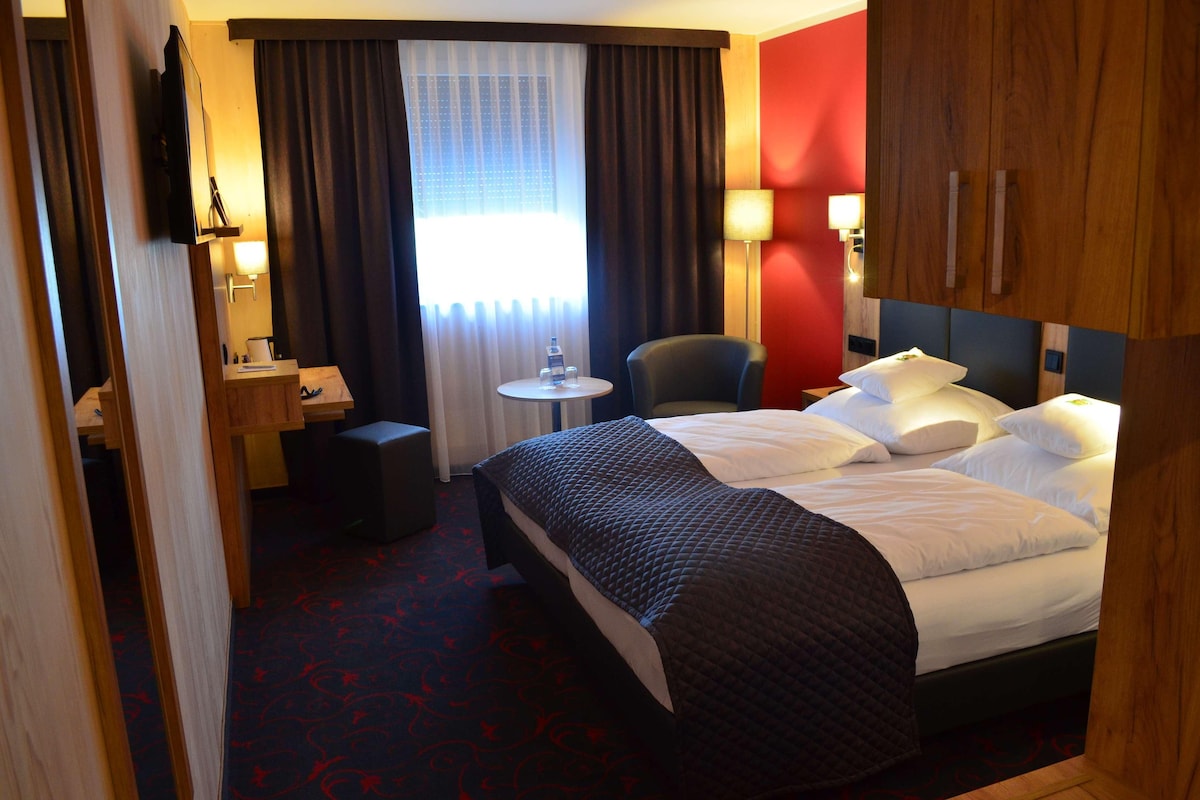 A hotel room with a double bed, a small desk, a chair, a round table, and a view of the city through the window.