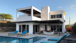 Una casa moderna de dos pisos con piscina, mobiliario de exterior y una hamaca.