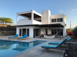 Una casa moderna de dos pisos con piscina, mobiliario de exterior y una hamaca.