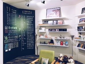 A modern interior with a chalkboard displaying event schedules, shelves with books and decorative items, and a green chair with a 'Good Luck' pillow.
