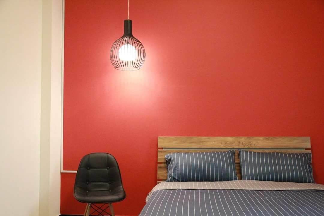Un dormitorio con una pared roja, un cabecero de madera, ropa de cama a rayas, una silla negra y una lámpara colgante.