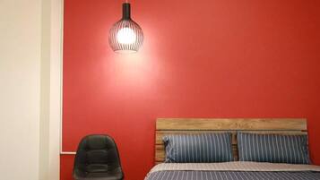 Un dormitorio con una pared roja, un cabecero de madera, ropa de cama a rayas, una silla negra y una lámpara colgante.