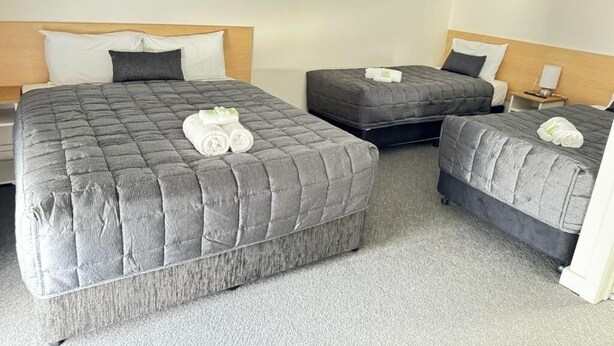 Habitación de hotel con tres camas, cada una con edredón y almohadas grises acolchadas. Se observa una mesita de noche con lámpara a un lado y una mesita auxiliar al otro.