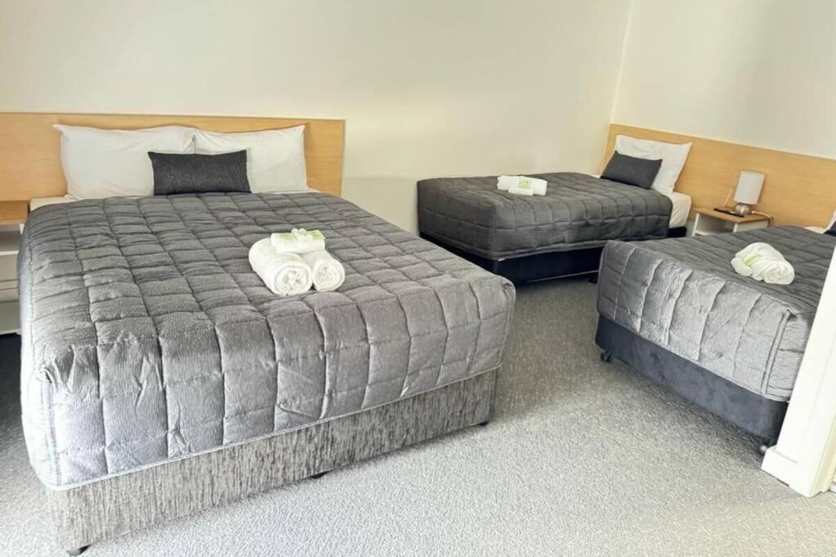 A hotel room with three beds, each with a grey quilted duvet and pillows. There is a small bedside table with a lamp on one side and a nightstand on the other.