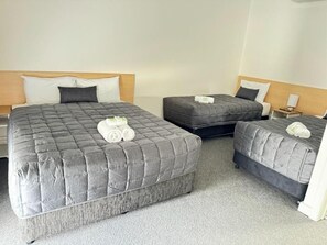 A hotel room with three beds, each with a grey quilted duvet and pillows. There is a small bedside table with a lamp on one side and a nightstand on the other. - Twin City Motor Inn (Wodonga)