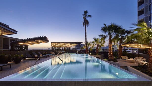 Una piscina infinita en la azotea con sillones de descanso y palmeras, rodeada de edificios modernos.