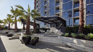 Un patio de hotel moderno con palmeras, mobiliario exterior y una zona cubierta con diseño de celosía.