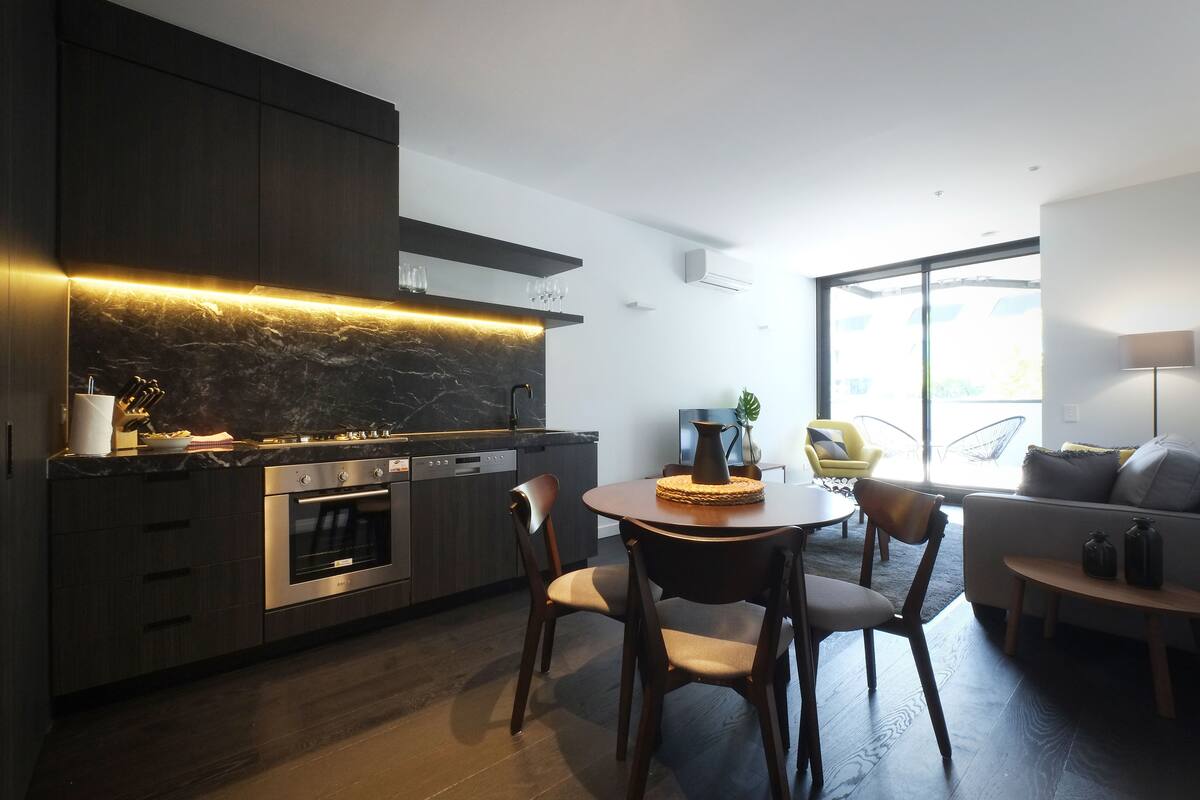 Una cocina moderna con armarios oscuros, horno integrado y un comedor con mesa y sillas redondas.
