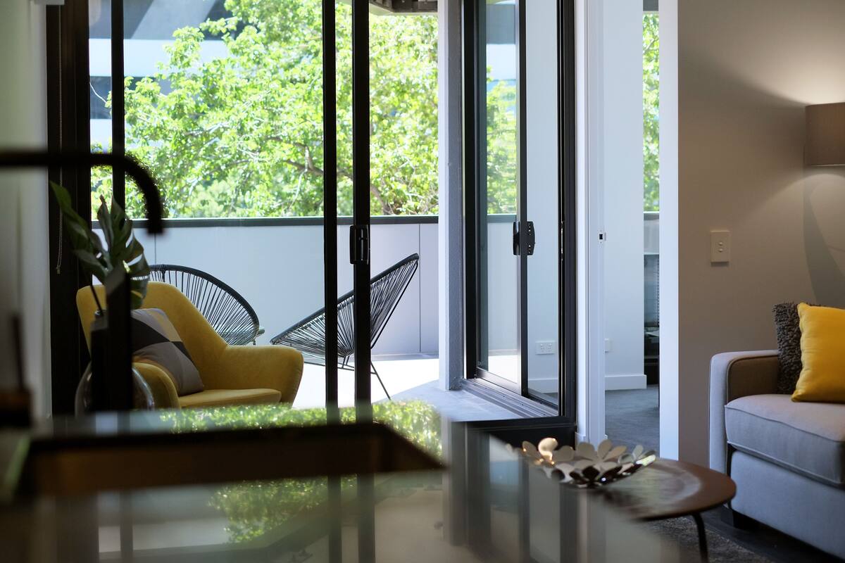 Una sala de estar moderna con un ventanal de vidrio que da a un balcón, un sofá, una silla y una mesita con adornos.