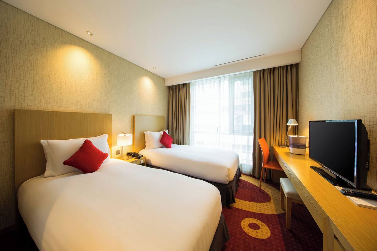 A hotel room with two beds, a desk, a TV, and a window with curtains.