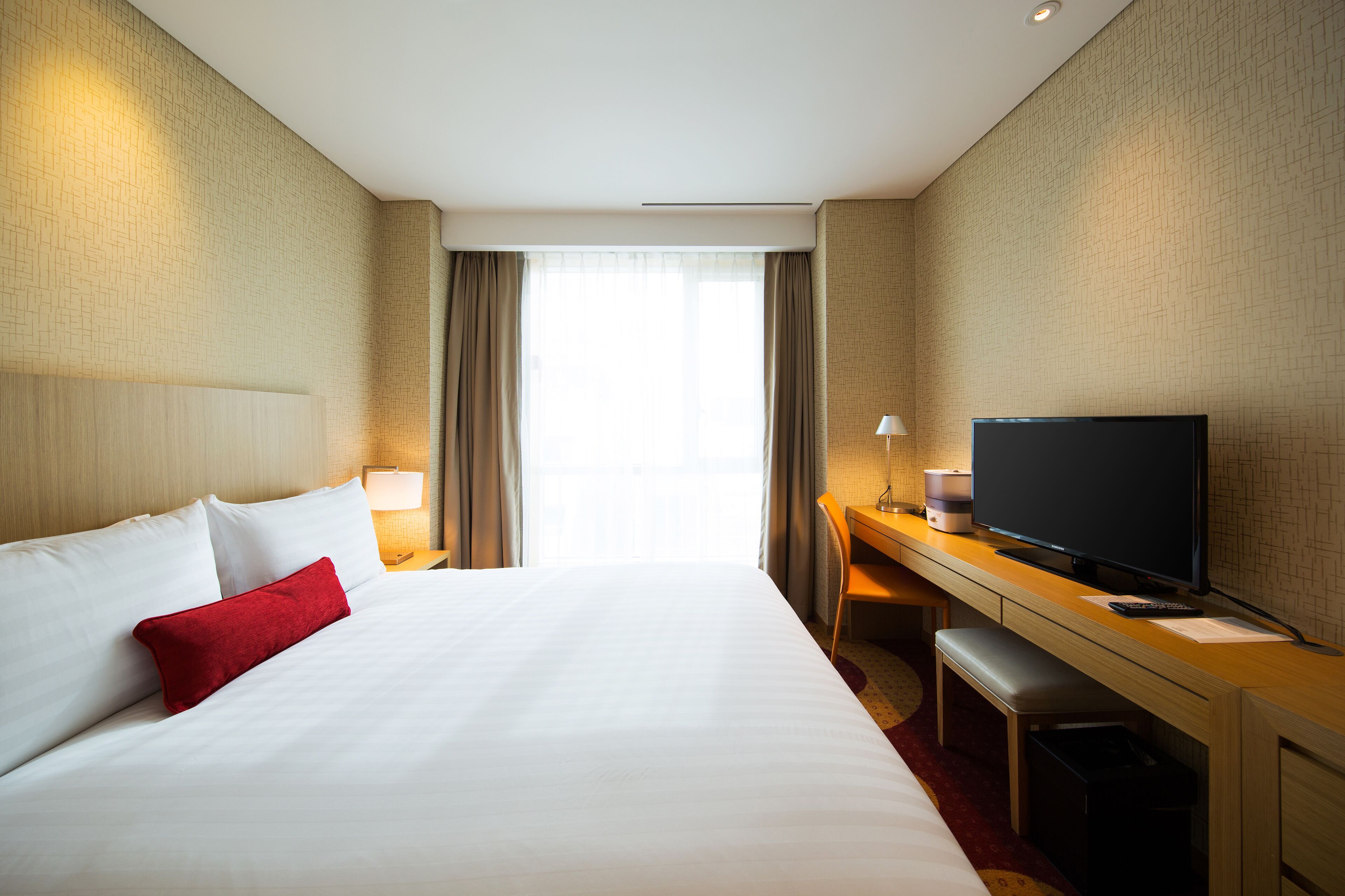 A hotel room with a large bed, a desk with a television, and a view of the city through the window.
