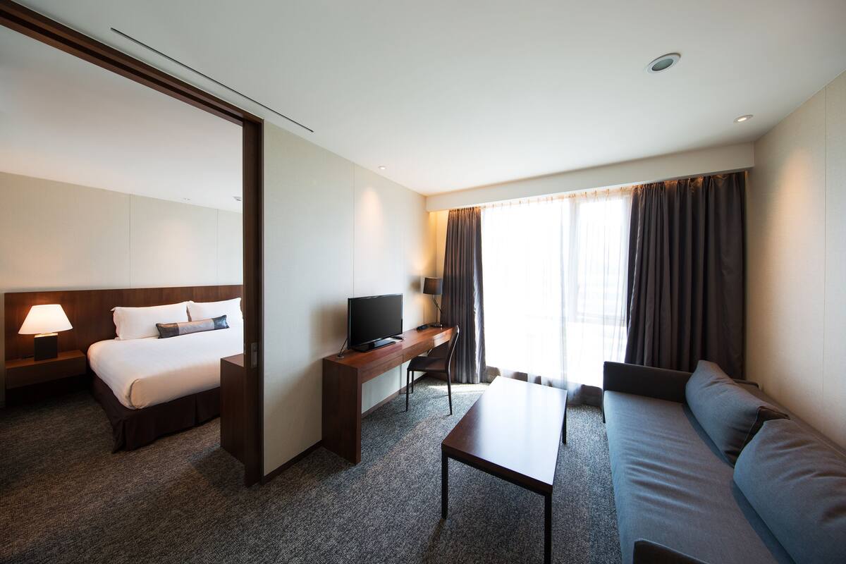 A hotel room with a bed, a desk with a television, a sofa, and a large window with curtains.