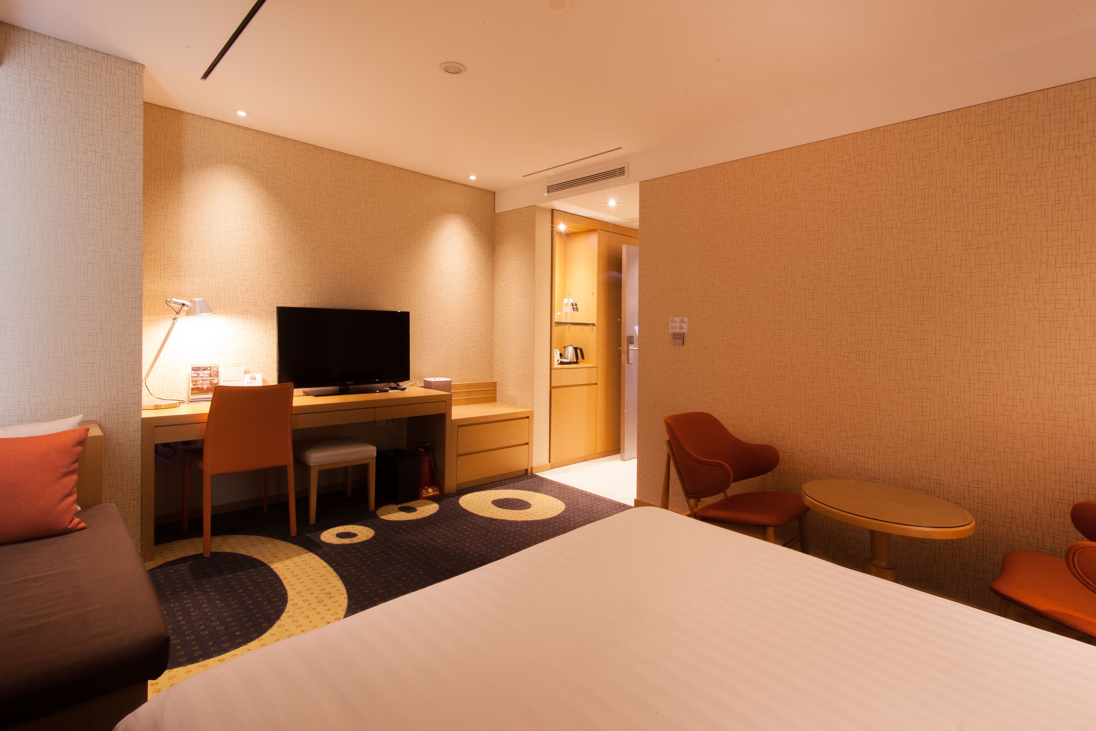 A hotel room with a bed, a desk with a computer, a TV, and a small table with chairs.