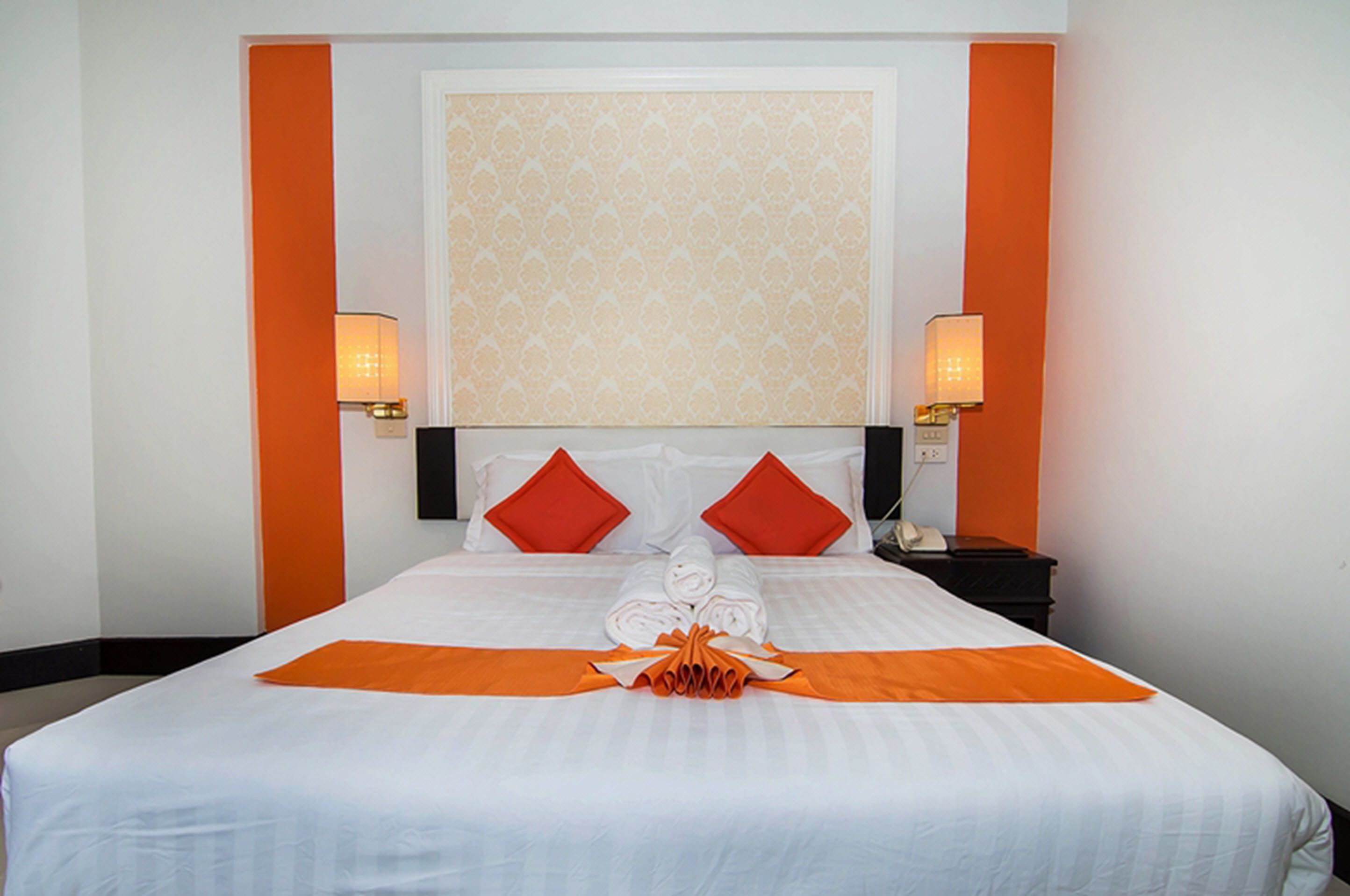 A hotel room with a neatly made bed, two red pillows, a folded towel, and a decorative orange bow.