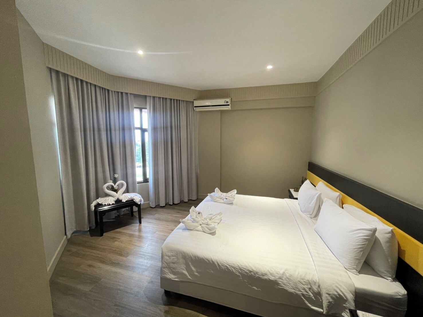 A hotel room with a large bed, a bedside table, a window with curtains, and an air conditioning unit.