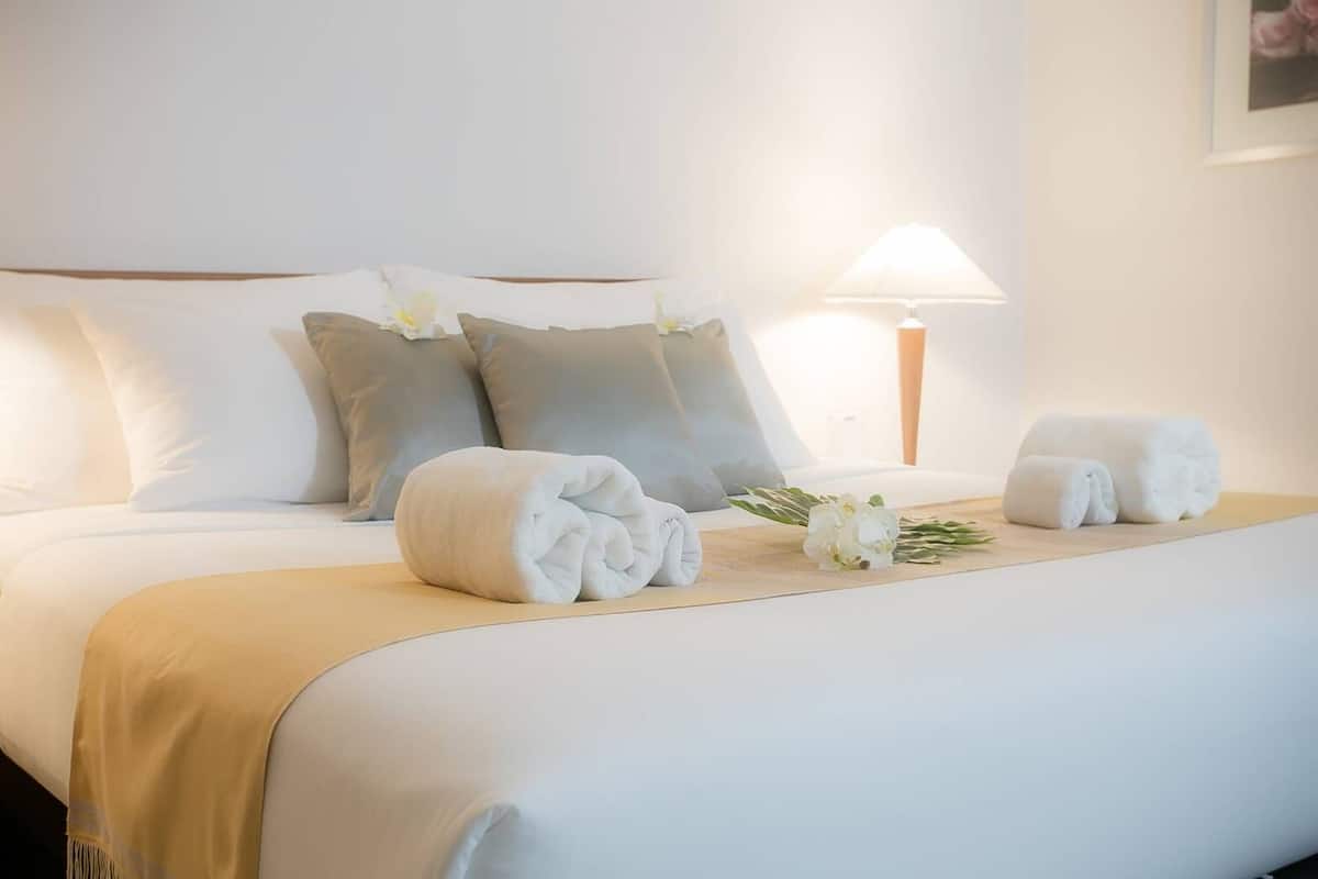 A neatly made bed with white and grey pillows, two rolled towels, and a small bouquet of white flowers.