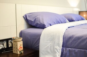 Una cama bien hecha con una colcha blanca y morada, un pequeño adorno en la mesita de noche y una foto en un marco en la pared.