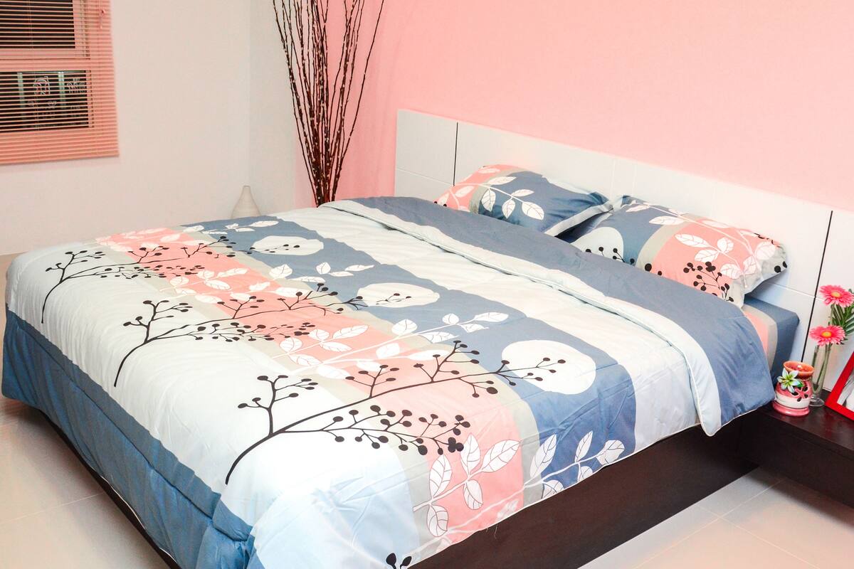 A neatly made bed with floral bedding, a bedside table with a picture frame and flowers, and a window with blinds.