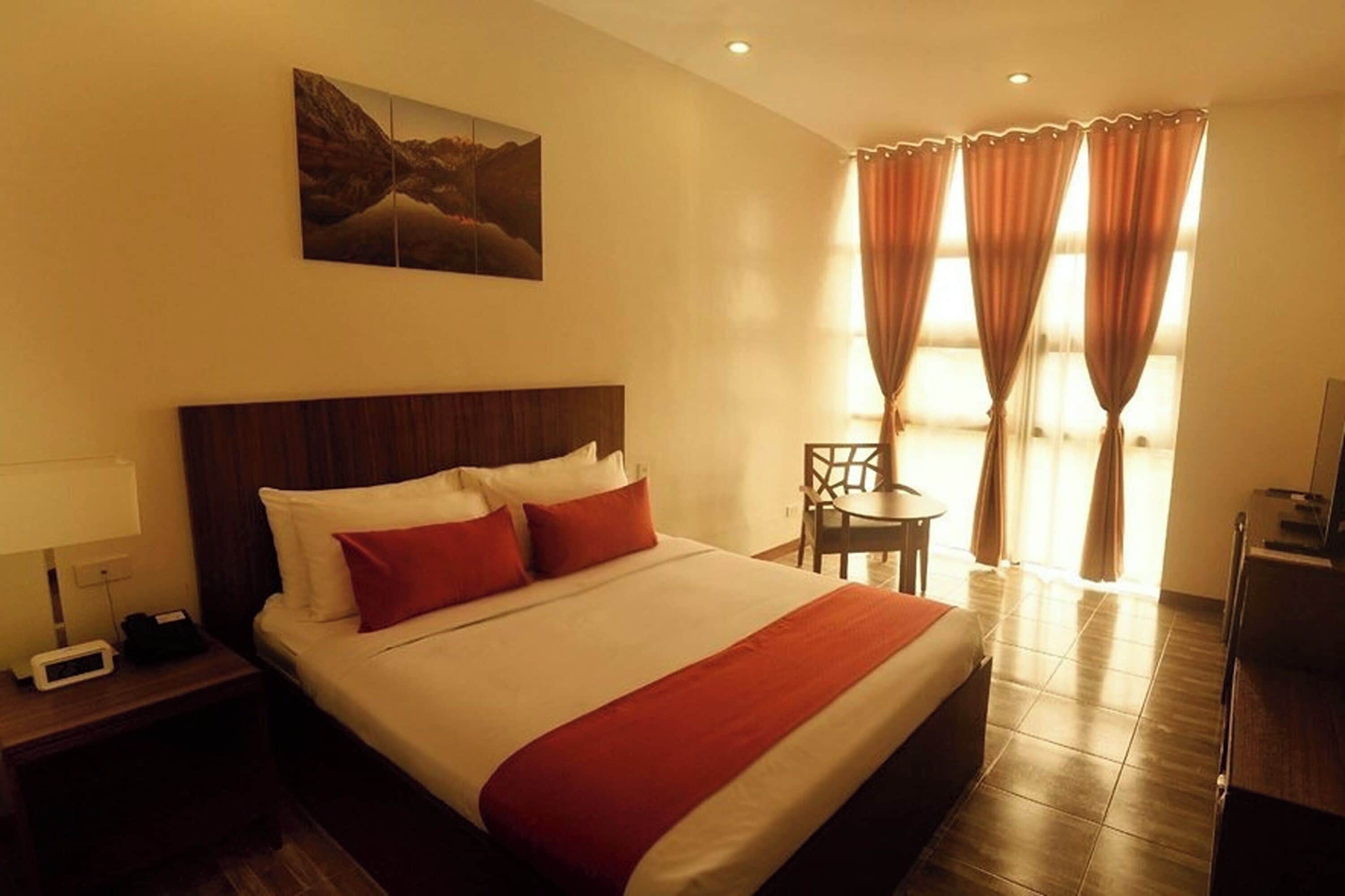 Quarto de hotel com uma cama grande, mesa de cabeceira com luminária, uma cadeira e uma escrivaninha.